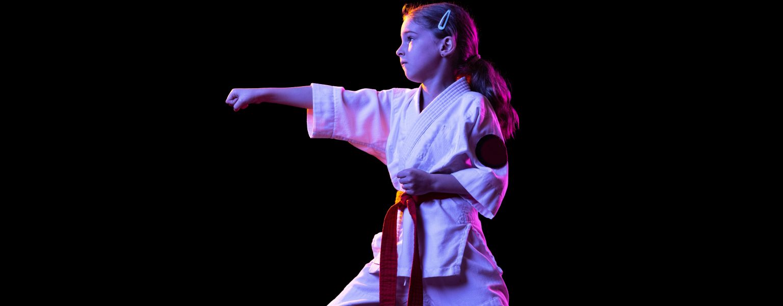 Portrait of little girl in white kimono training martial art isolated over black background in neon